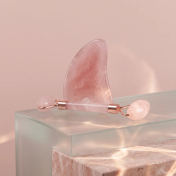 Gua sha et rouleau de massage rose posés sur un bloc de verre