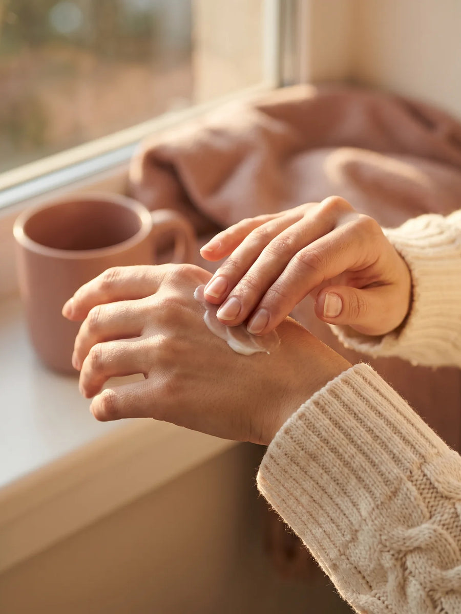 Femme qui applique de la crème sur ses mains sèches, au coin d'une fenêtre