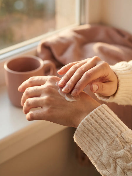 Femme qui applique de la crème sur ses mains sèches, au coin d'une fenêtre